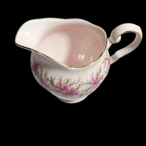 May include: A vintage pink creamer with a floral design. The creamer has a curved handle and a gold trim around the rim. Pink flowers and green leaves decorate the exterior. The creamer is made of porcelain.