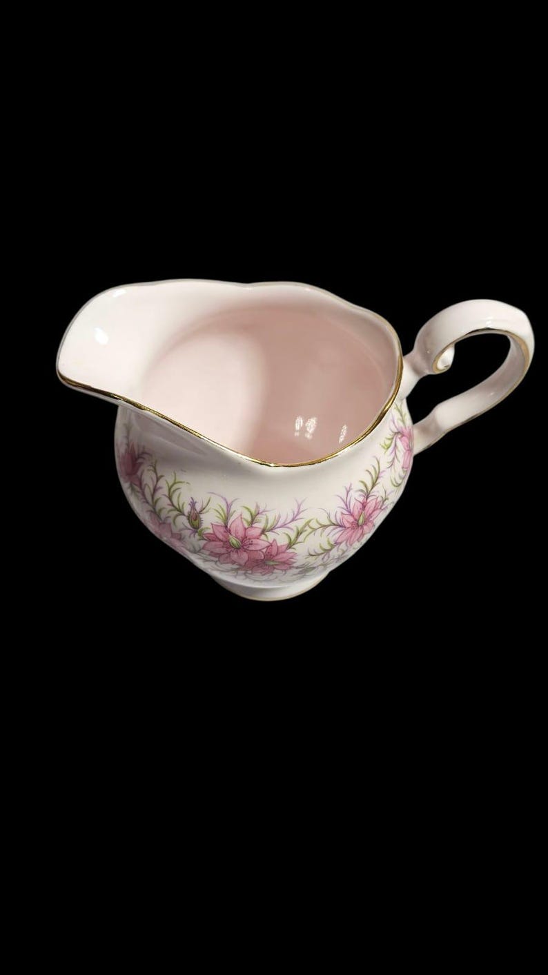 May include: A vintage pink creamer with a floral design. The creamer has a curved handle and a gold trim around the rim. Pink flowers and green leaves decorate the exterior. The creamer is made of porcelain.