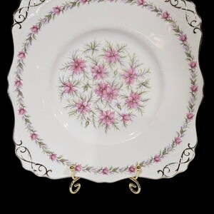 May include: A square, white ceramic plate with a floral design. The plate features a border of pink flowers and green leaves, with a central cluster of pink flowers. Gold accents adorn the scalloped edges.