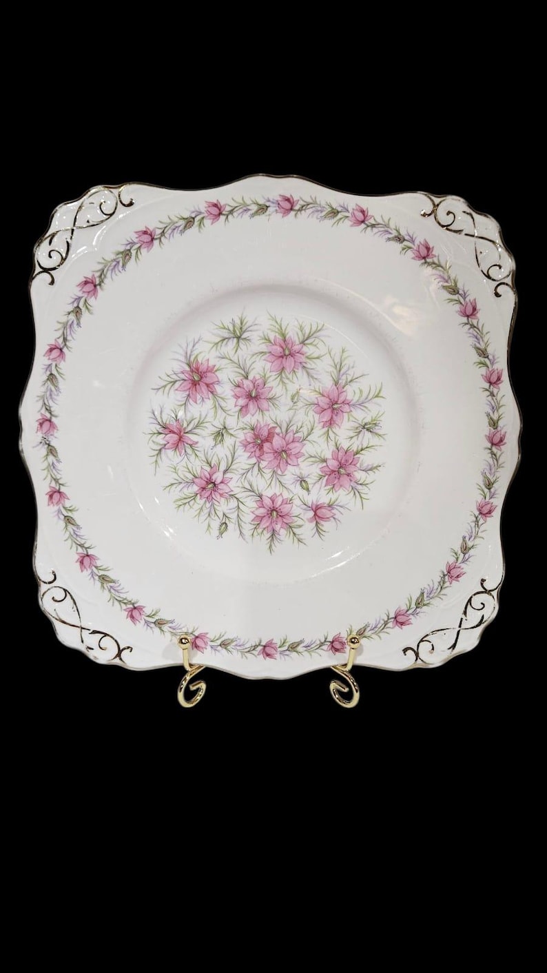 May include: A square, white ceramic plate with a floral design. The plate features a border of pink flowers and green leaves, with a central cluster of pink flowers. Gold accents adorn the scalloped edges.
