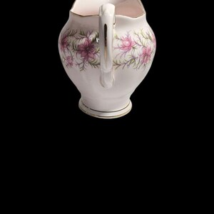May include: A white ceramic creamer with a handle and a floral design featuring pink flowers and green leaves. The creamer has a gold trim around the rim and base. The creamer is approximately 5 inches tall.