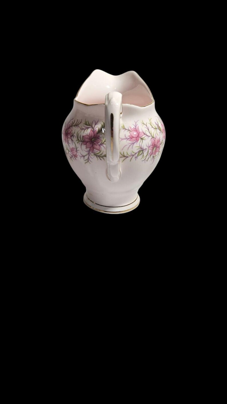 May include: A white ceramic creamer with a handle and a floral design featuring pink flowers and green leaves. The creamer has a gold trim around the rim and base. The creamer is approximately 5 inches tall.