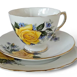May include: A white teacup, saucer, and plate set. The teacup and plates are decorated with yellow roses, blue flowers, and green leaves. The edges of the teacup and plates are trimmed with gold.