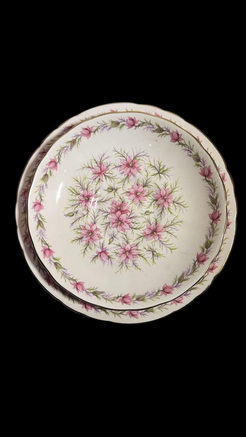 May include: Two white ceramic plates with a floral design. The plates feature a pink floral pattern in the center and around the rim, with a gold trim. The plates are set against a black background.