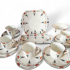 Może przedstawiać: Biały porcelanowy zestaw do herbaty z kwiatowym wzorem. Zestaw zawiera filiżanki, spodki, półmisek, miskę i prostokątny talerz. Kwiatowy wzór ma czerwone i czarne akcenty. Styl vintage.