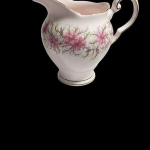 May include: A white ceramic creamer with a floral pattern of pink flowers and green leaves. The creamer has a gold trim around the rim, base, and handle. The handle is a curved, elegant design.