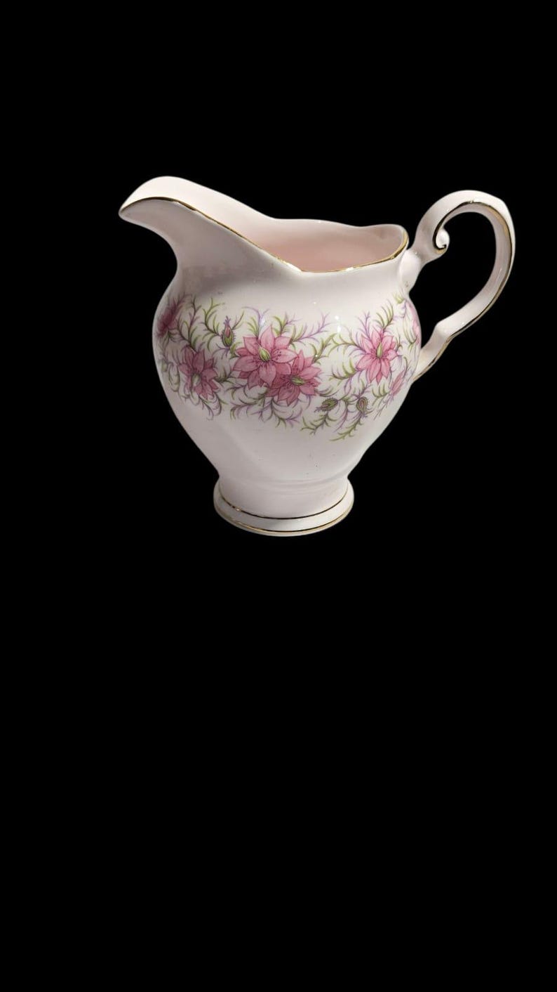 May include: A white ceramic creamer with a floral pattern of pink flowers and green leaves. The creamer has a gold trim around the rim, base, and handle. The handle is a curved, elegant design.