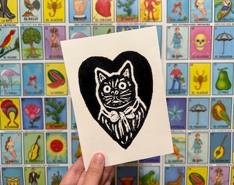 May include: A black and white linocut print of a cat inside a heart shape. The cat has wide eyes and a bow tie. The background features a colorful wall of Mexican Loteria cards.