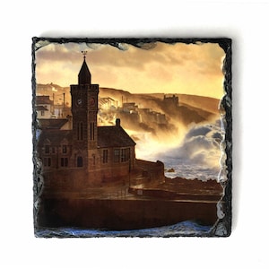 May include: A photograph of a town with a clock tower in the foreground. The town is situated on a cliff overlooking a stormy sea. The sky is a vibrant orange and yellow, and the waves are crashing against the rocks.