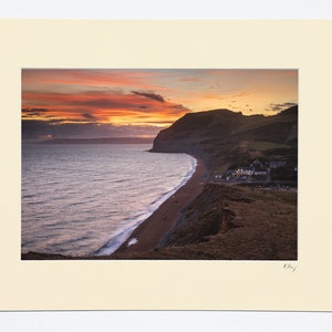 May include: A scenic photograph of a coastal town at sunset. The image shows a long, sandy beach with waves crashing on the shore. A small town is nestled at the base of a cliff, with a dramatic sky filled with vibrant orange, pink, and purple hues.