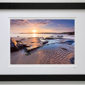 May include: A photograph of a beach at sunset with the sun setting over the water. The sand is wet and there are rocks and tide pools in the foreground. The sky is a mix of pink, purple, and orange.