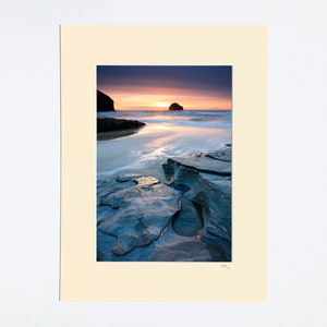 May include: A photograph of a rocky beach at sunset with a small island in the distance. The water is calm and reflects the colours of the sky, which is a mix of purple, pink, and orange. The rocks are a dark grey colour and are covered in water.