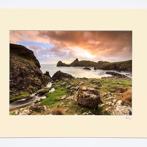 May include: A scenic photograph of a rocky coastline with a small stream flowing through the foreground. The sky is a vibrant orange and pink, with clouds casting shadows on the water.