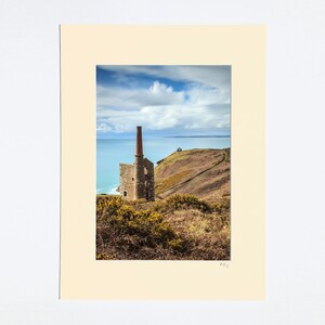 May include: A ruined stone building with a tall red brick chimney stands on a grassy hillside overlooking a blue sea. The building is partially derelict, with some of the walls missing. The sky is a bright blue with white clouds.