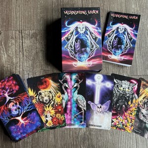May include: A tarot deck with a black background and colorful illustrations. The box features a woman with flowing hair and a glowing orb. The cards depict various scenes with animals, plants, and mystical symbols. The deck is called "Vespertine Tarot".