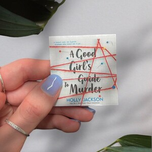 以下が含まれることがあります： 赤と白のデザインのペーパーバックの表紙。タイトルは「A Good Girl's Guide to Murder」で、作者はホリー・ジャクソン。この本はニューヨーク・タイムズのベストセラーです。
