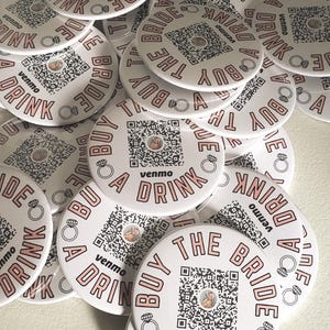 Puede incluir: Posavasos circulares blancos con texto rosa que dice "Buy the Bride a Drink" y un código QR. Los posavasos también presentan un gráfico de anillo y el logotipo de Venmo. Se muestra una pila de estos posavasos.