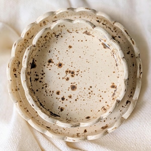 May include: A stack of three off-white ceramic dishes with a scalloped edge design. The dishes have a speckled pattern of brown spots. The plates are nested together, showcasing their varying sizes and textures. The background is a soft, white fabric.
