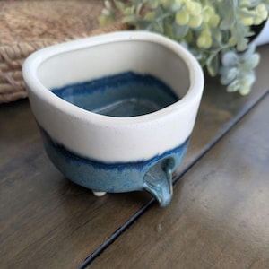 May include: A small, white ceramic planter with a blue glaze. The planter has a rounded, rectangular shape and three small feet. It has a small spout on one side.
