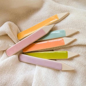 May include: Six ceramic plant markers in various pastel colors, including pink, orange, green, and yellow. The markers are shaped like long, thin rectangles with pointed ends.