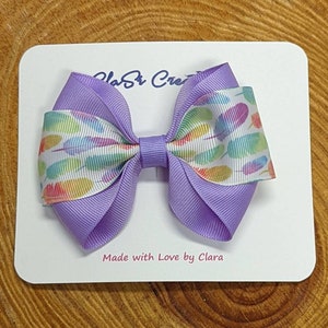 May include: A purple hair bow with a white background and a colorful feather print. The bow is on a white card with the text "Made with Love by Clara".