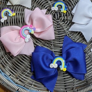 May include: Six hair bows with rainbow and cloud embellishments. The bows are in various colors, including white, light blue, pink, and dark blue. The bows are arranged in a wicker basket.
