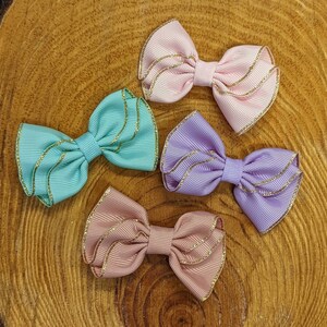 May include: Four handmade hair bows in pink, light blue, lavender, and a darker shade of pink. Each bow has a gold glitter trim and is made of a soft, textured fabric.