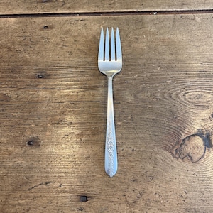 May include: A silver fork with four tines, resting on a rustic wooden surface. The handle features an ornate floral design. This utensil is suitable for everyday dining.