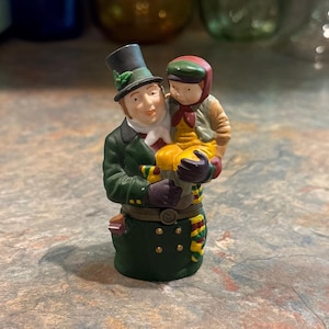 May include: A ceramic figurine of a man in a green coat and top hat holding a child in a yellow and red outfit. The man has a white beard and the child is wearing a red hat.