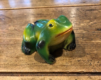 May include: A green frog figurine with black spots, sitting on a wooden surface. The frog has a yellow eye and an open mouth.