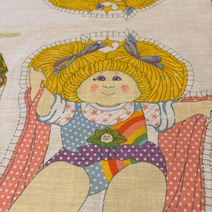 May include: A vintage fabric panel featuring a cartoon illustration of a Cabbage Patch Kid doll wearing a rainbow and star-patterned outfit. The doll has blonde hair, blue eyes, and a pink blush. The fabric panel is ideal for crafting projects such as quilts, cushions, and tote bags.