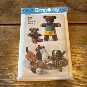 Może przedstawiać: Wzorzec szycia vintage Simplicity 9647 do zestawu pluszowych zwierzątek. Wzór zawiera instrukcje do uszycia brązowego misia, czarno-białego psa w kropki i psa ze szalikiem i czapką w kratę.