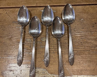 May include: Five silver spoons with ornate floral designs on the handles. The spoons are arranged in a row on a wooden surface.