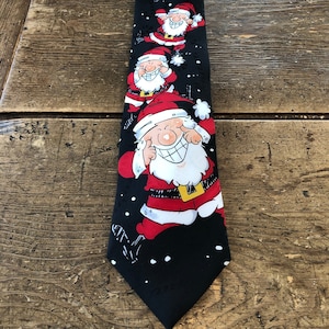 Może przedstawiać: Czarny krawat z powtarzającym się wzorem postaci Świętego Mikołaja w kolorze czerwonym, białym i czarnym. Postacie Świętego Mikołaja uśmiechają się i mają białe brody i wąsy. Tło jest czarne z białymi płatkami śniegu.