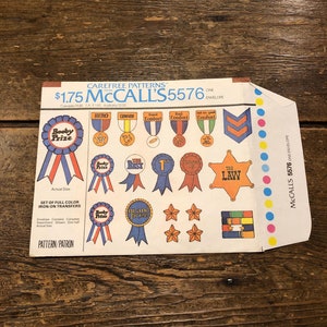 Puede incluir: Un conjunto de transferencias de planchado con cintas de colores, estrellas y insignias con frases como "Body Prize", "The Best", "Grand Prize" y "The Law". El paquete está etiquetado como "McCall's 5576" e incluye instrucciones de uso.