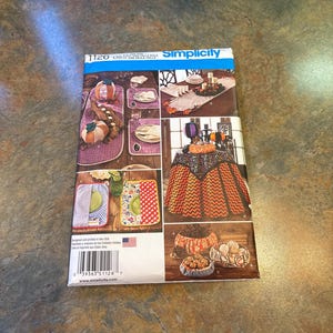 Op de afbeelding: Simplicity naaipatroon nummer 1126 voor een tafelrok, placemats en pompoen-thema tafeldecoraties. Het patroon bevat instructies voor het maken van een tafelrok met een zwart en oranje chevron-patroon, placemats met verschillende patronen en pompoen-vormige decoraties. Het patroon is ontworpen en gedrukt in de VS.