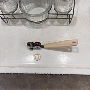 May include: A manual knife sharpener with a beige plastic handle and two metal sharpening wheels, positioned next to a United States quarter coin for scale. The sharpener features a metallic frame and a small hole at the end of the handle.