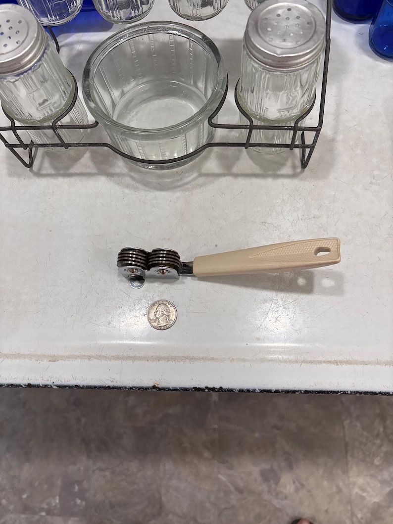 May include: A manual knife sharpener with a beige plastic handle and two metal sharpening wheels, positioned next to a United States quarter coin for scale. The sharpener features a metallic frame and a small hole at the end of the handle.