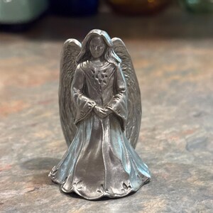 May include: A small, silver-colored angel figurine with outstretched wings. The angel is wearing a long gown and has its hands clasped in front. The figurine is detailed and stands on a textured surface.