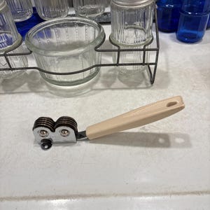 May include: A handheld knife sharpener featuring a beige plastic handle with a textured grip and a hole at the end. The sharpening mechanism consists of two dark brown circular wheels held within a chrome-plated metal frame marked with the text EKCO.