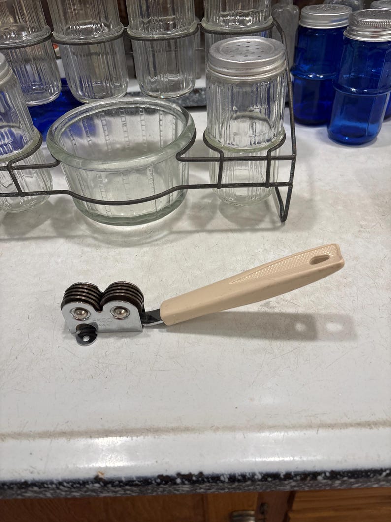 May include: A handheld knife sharpener featuring a beige plastic handle with a textured grip and a hole at the end. The sharpening mechanism consists of two dark brown circular wheels held within a chrome-plated metal frame marked with the text EKCO.