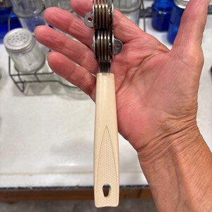 May include: A manual knife sharpener featuring a cream-colored plastic handle with a textured diamond pattern and a small hanging hole at the base. The sharpening head consists of two stacked metal wheels held within a silver-toned metal frame.