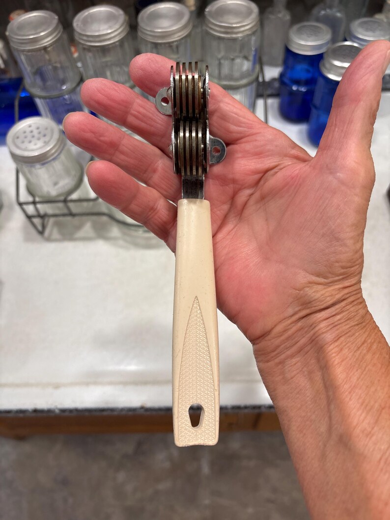 May include: A manual knife sharpener featuring a cream-colored plastic handle with a textured diamond pattern and a small hanging hole at the base. The sharpening head consists of two stacked metal wheels held within a silver-toned metal frame.