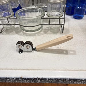 May include: A handheld knife sharpener featuring a beige plastic handle and a chrome-plated metal head with two dark brown sharpening wheels. The metal head is stamped with the brand name EKCO and the letters USA, indicating its origin and manufacturer.