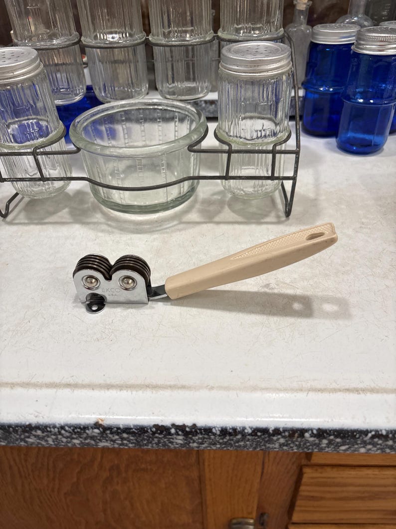May include: A handheld knife sharpener featuring a beige plastic handle and a chrome-plated metal head with two dark brown sharpening wheels. The metal head is stamped with the brand name EKCO and the letters USA, indicating its origin and manufacturer.