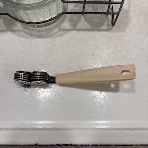 May include: A kitchen knife sharpener featuring a beige plastic handle with a textured triangular design and a hole at the end. The sharpening mechanism consists of two sets of stacked, circular metal discs held together by a metal frame.