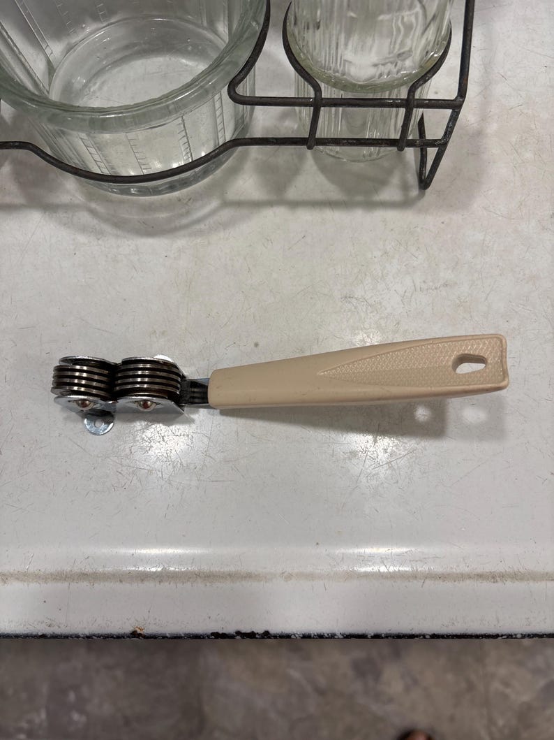 May include: A kitchen knife sharpener featuring a beige plastic handle with a textured triangular design and a hole at the end. The sharpening mechanism consists of two sets of stacked, circular metal discs held together by a metal frame.