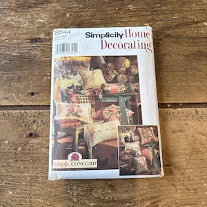 Puede incluir: Un libro de patrones vintage de Simplicity Home Decorating. La portada presenta un collage de cojines decorativos y el texto "Simplicity Home Decorating". El libro está sobre una superficie de madera.