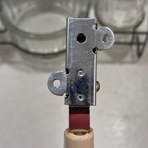 May include: A manual can opener featuring a rectangular silver-colored metal head with two circular side tabs and two small rivets. The head is attached to a solid, light beige cylindrical handle by a flat, dark red metal connector piece.