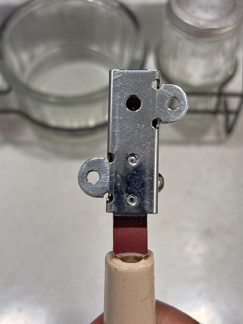 May include: A manual can opener featuring a rectangular silver-colored metal head with two circular side tabs and two small rivets. The head is attached to a solid, light beige cylindrical handle by a flat, dark red metal connector piece.